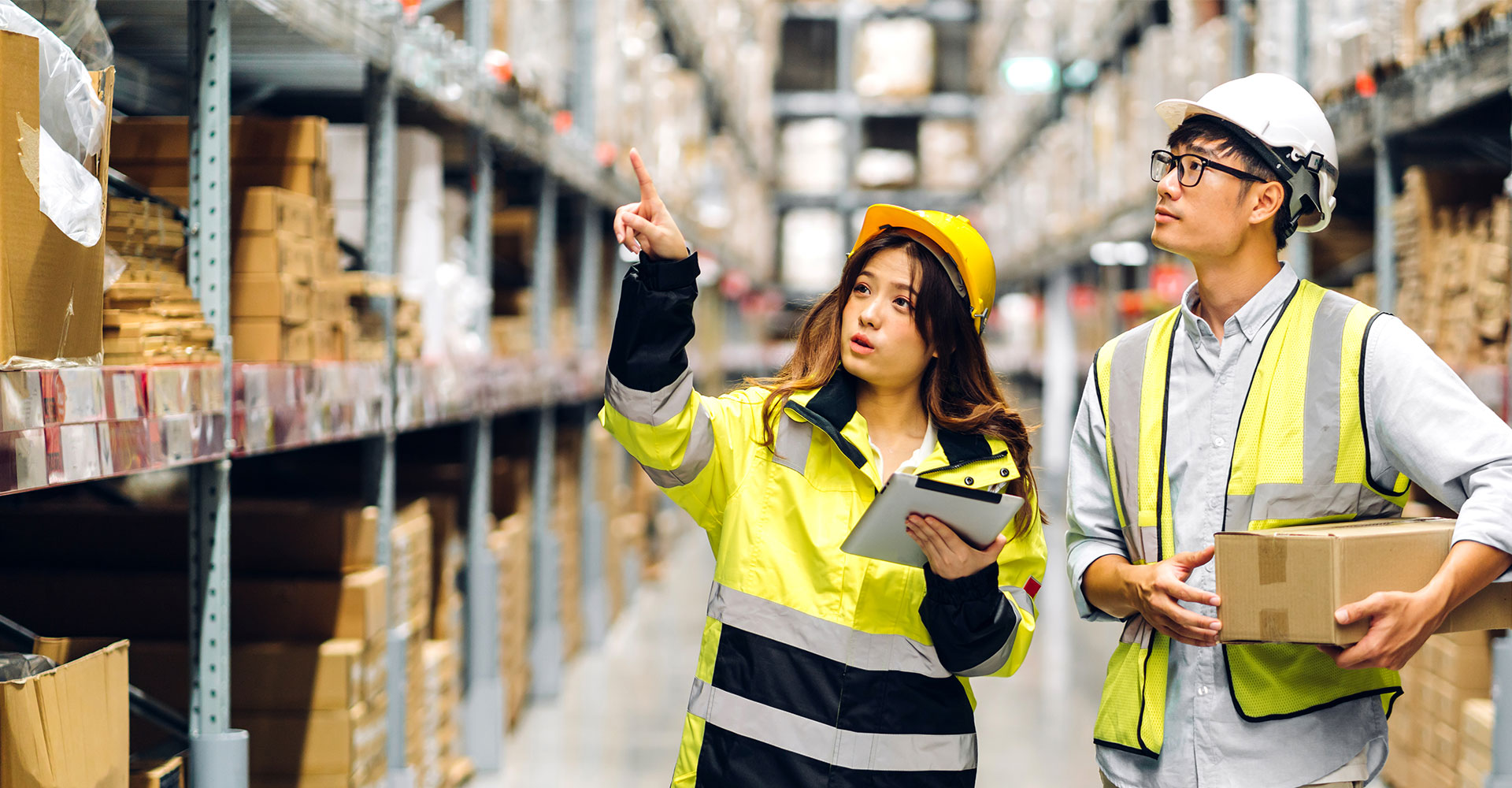 Young female supply chain assistant in high viz overcoat and safety gear coordinating with supply chain manager in hard hat on current inventory needs