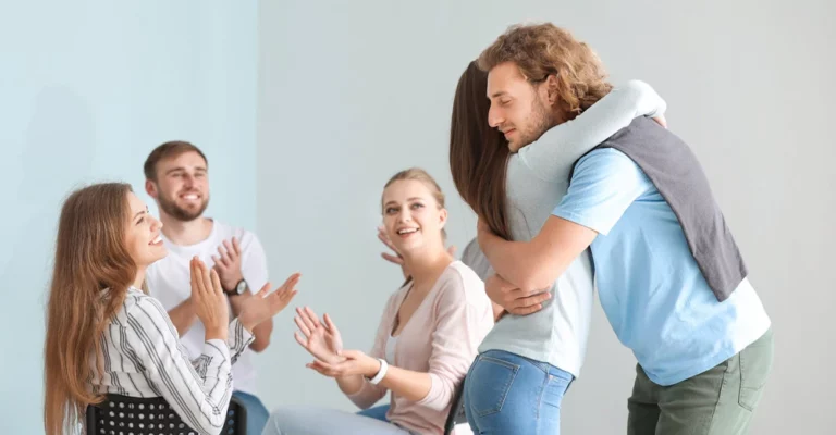 In a room, a young man struggling with mental health issues hugs a supportive woman while the rest of the support group applauds, acknowledging his openness in sharing his experiences.