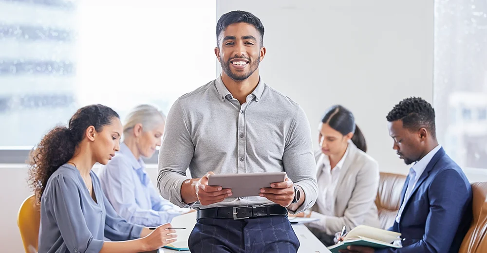 An entrepreneur with a tablet leans against a table as his team collaborates on business strategies.