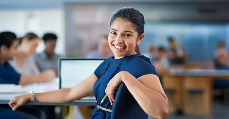 Portrait of a South Asian woman with her hair pulled into a low ponytail with a pencil in hand, smiling confidently in a post-secondary classroom while attending a career-focused course. Her laptop and other students are visible in the blurred background, engaged in learning.