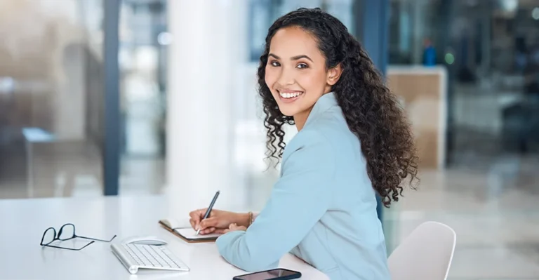 A young professional woman smiling at a desk with glasses, a keyboard, mouse, and phone while writing in notebook, searching for the best skills to put on a resume.