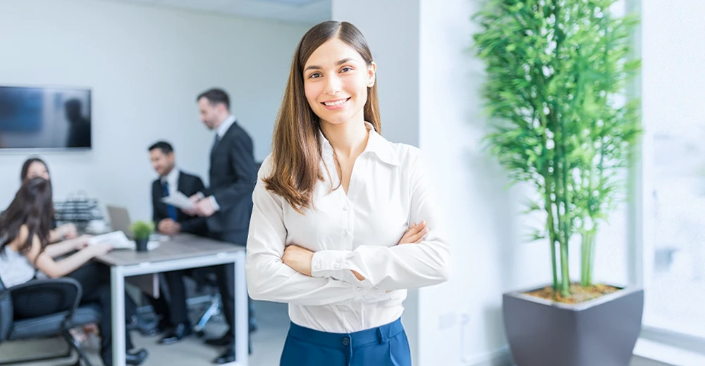 Smiling HR coordinator in a modern office with a team meeting in the background.