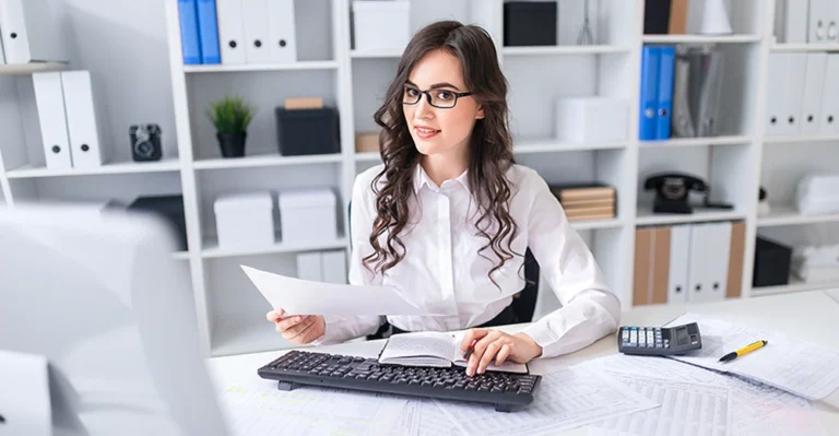Woman sitting behind a computer desk glancing to the right with paperwork and a calculator.