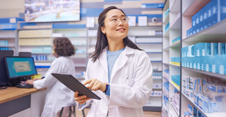 A woman in a white coat using a tablet in pharmacy beside shelves of medicine.